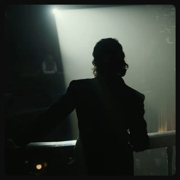 Man with dark, wavy, shoulder-length hair; wearing a suit; leaning against a theatre balcony with his back to the camera. A bright spotlight illuminates his silhouette. 